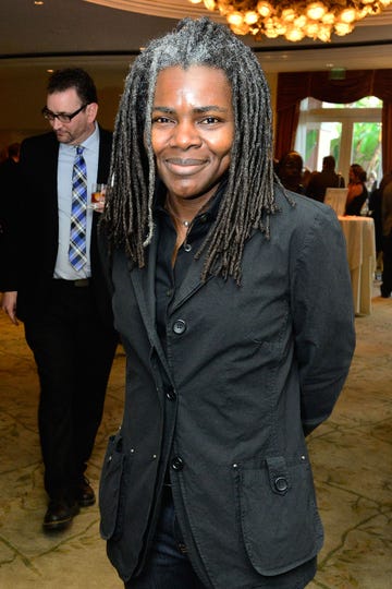 tracy chapman smiles at the camera while standing inside an event space with a chandelier, she wears a black jacket and black collared shirt, her dreads are slightly gray at the roots and reach past her shoulders