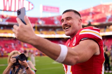 travis kelce smiles as he holds a phone out in front of his face while standing inside a football stadium on the field, he wears a red kansas city chiefs jersey