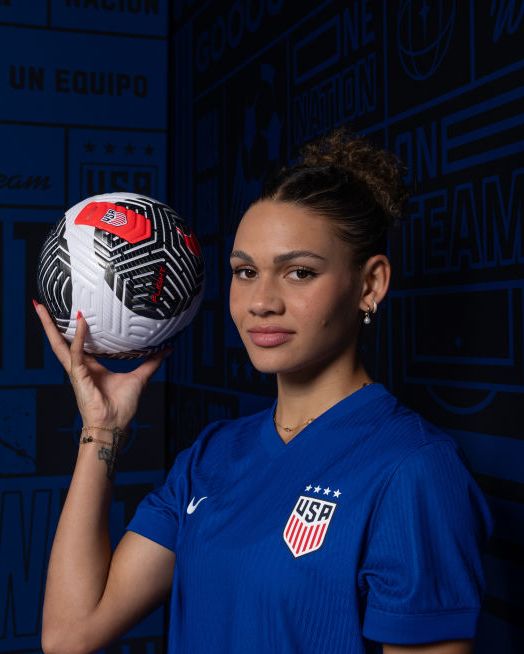 trinity rodman wearing her blue us soccer jersrey and holding a soccer ball in her right hand