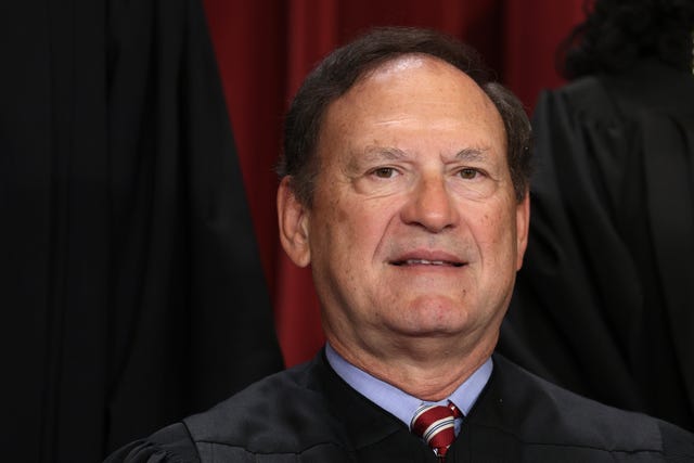 samuel alito smiles slightly at the camera, he wears black robes over a light blue collared shirt and red tie with a white and blue stripe