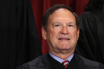 samuel alito smiles slightly at the camera, he wears black robes over a light blue collared shirt and red tie with a white and blue stripe