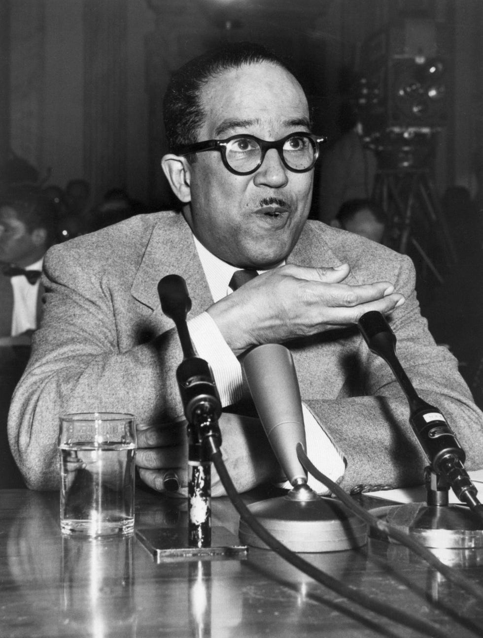 langston hughes gesturing with his right hand as he sits in front of a desk with microphones on it and speaks