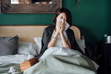 young asian woman sitting on bed and blowing her nose with tissue while suffering from a cold, with medicine bottle and a glass of water on the side table