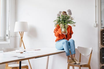 young woman sitting on table holding flower vase in front of her face