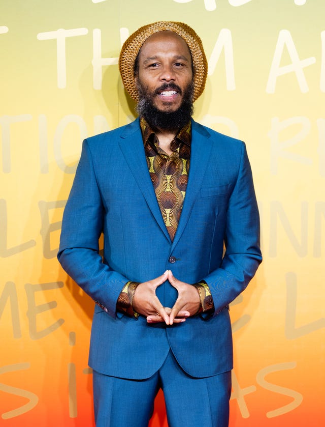 ziggy marley smiles while standing in front of a green, yellow and orange photo background, he wears a blue suit with a green and brown patterned shirt and brown knit cap