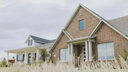 A brick house and a white house next door to each other with tall grass in the foreground.