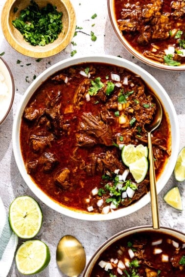 Bowl of birria served with diced onion and cilantro with a lime wedge.