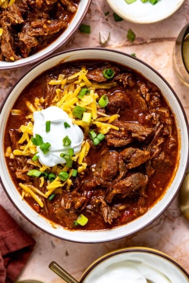 A bowl of hearty chuck roast chili topped with shredded cheese, sour cream, and chopped green onions.