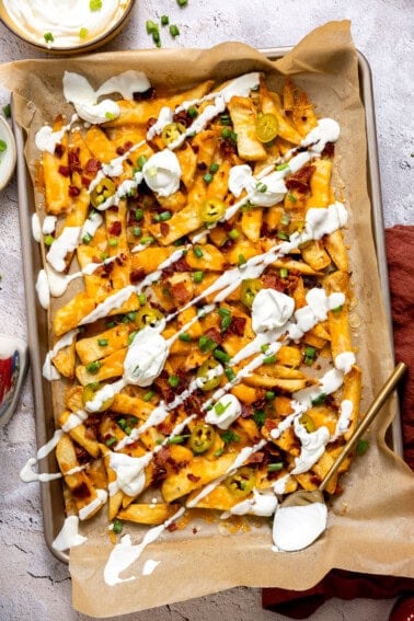 Golden, crispy French fries loaded with melted cheese, bacon, green onions, and a drizzle of sour cream on a baking tray.