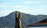 Foto de uma estátua apontando para o céu. Um pouco atrás, o telhado de uma casa e, ao fundo, colinas cobertas por vegetação.