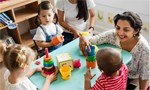 Três crianças sentadas em torno de uma mesa com alguns brinquedos. Duas adultas supervisionam as atividades: uma, parcialmente fora do enquadramento, e a outra segura um brinquedo enquanto interage com uma das crianças. Mais ao fundo, há uma prateleira de madeira com livros e alguns desenhos pendurados na parede.