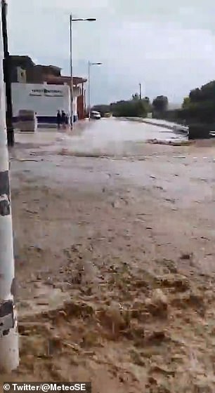 Water is seen pouring down a street and over a junction before crashing into a small barrier