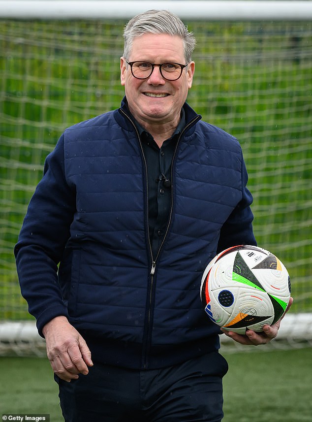 With a smart navy puffer jacket with fabric sleeves, Keir Starmer looks pleased as he holds a football