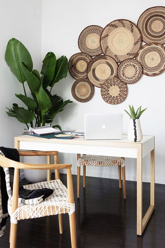 A boho tropical home office with a sleek desk, woven chairs, potted plants, decorative plates on the wall is very airy and cool.