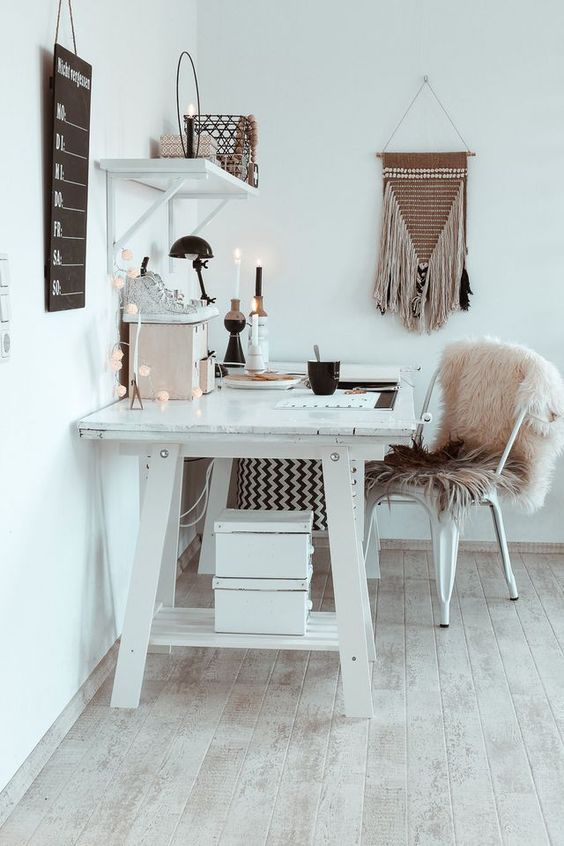 A white boho home office with a large desk, a metal chair with a pink fur cover, candles, lights and a macrame hanging.