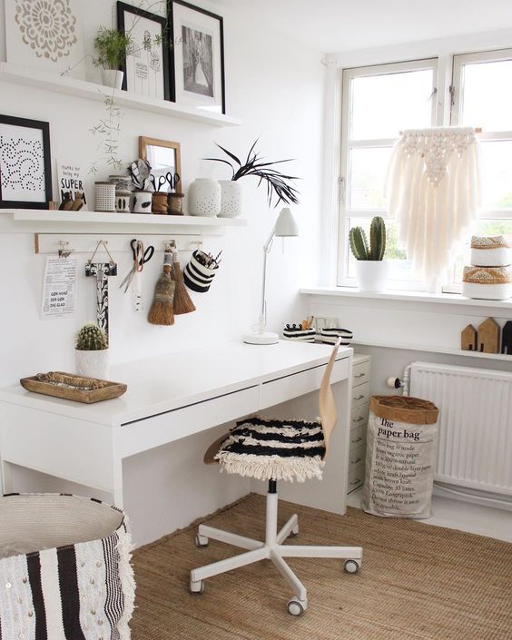 A white boho home office with open shelves, a white desk, a chair with a fringe cover, a boho ottoman, a macrame hanging and potted plants.