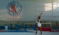 Biles shown in gymnastics floor pose in a training hall. On the wall is a huge banner that reads: 'The World Is Yours!'