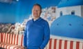 Donat Retif poses for a photo with his hands in his pockets in front of a wall with a photo of a holiday destination