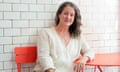 Middle-aged white women with long gray hair and white top sits at red metal bistro table along wall of white subway tiles, smiling for portrait.