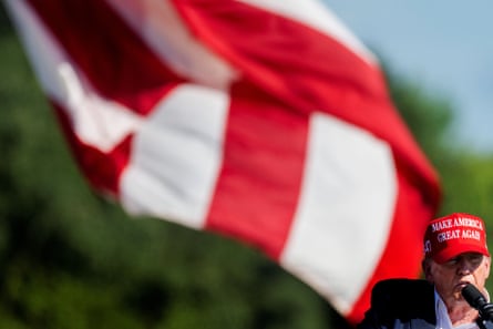 a man in a red Maga hat speaks in front of a billowing American flag