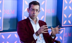 a man wearing a red jacket speaks while seated on stage