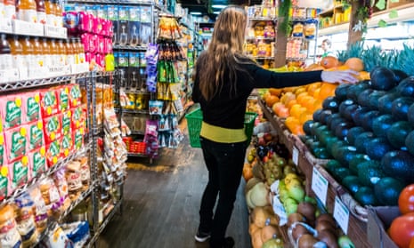 Seen from behind, a white woman with long blond hair reaches out a hand to select an avocado at a grocery store.