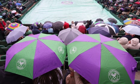 Two people hold umbrellas while sat in the seats at a tennis court