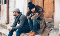 Young couple having relationship difficulties. Man and woman sitting on stairs in front of a closed wooden door. Wears warm clothes - jeans, sweater, coat, hat and scarf. Both looking in different directions. On background building facade, wooden front door, rainwater gutter and windows.