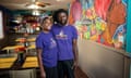 A Black couple wearing head wraps and purple t-shirts that say 'Afrodisiac' pose together in a restaurant that has a colorful wall mural featuring Black women and Jamaican Creole food ingredients