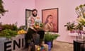 A man sits in a pink colored space surrounded by flowers and a portrait of a woman on the wall.