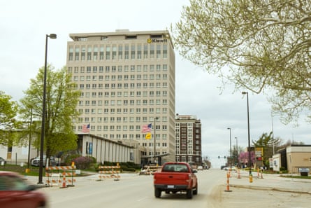 A maybe-10-story building in a sleepy-looking downtown, with a red pick-up truck driving under a cloudy sky.