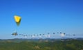 A tiny aircraft with two human passengers, a huge fan-like propeller on the back and a yellow parachute above leads a wavy line of birds against a clear blue sky.