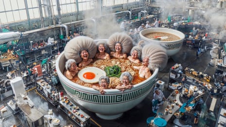 In a factory, laughing old Asian women sit in a giant bowl of ramen soup that resembles a hot tub