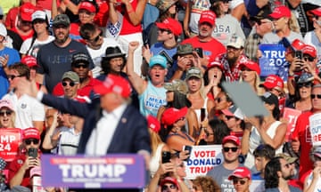 A frame full of supporters wearing red hats and holding signs, in front of an older man wearing a red baseball hat gesturing behind a podium.