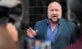 Alex Jones, with a beard and wearing a suit jacket and shirt unbuttoned by a few buttons, holds up his hands as he speaks in front of microphones