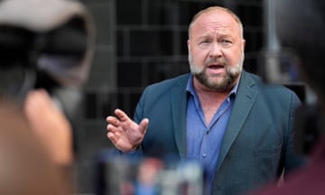 Alex Jones, with a beard and wearing a suit jacket and shirt unbuttoned by a few buttons, holds up his hands as he speaks in front of microphones