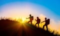 Three people walk up a mountain, with the sun setting in the background