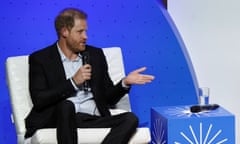 Prince Harry is seated on a white chair, speaking into a microphone, against a bright blue backdrop on a stage