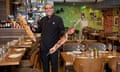 A smiling man stands in a dining room while holding meat on rotisserie skewers