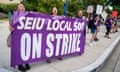 Three people stand on a sidewalk holding a large purple sign reading 'SEIU LOCAL 500 ON STRIKE' as several others hold signs nearby