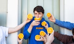 A man wearing a blue hoodie drinking from a glass of orange juice while people hold orange halves around his face.