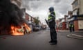 A police officer in riot gear looks at a car on fire outside a row of shops