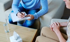 Two people sit on chairs next to a coffee table. One person is making notes on a clipboard