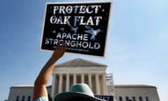 A person holds a sign reading 'Protect Oak Flat / Apache Stronghold' in front of a cream arched building