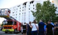 People gathered in front of the white apartment building, which has blackened windows in the middle on the top floor. A fire engine is parked alongside