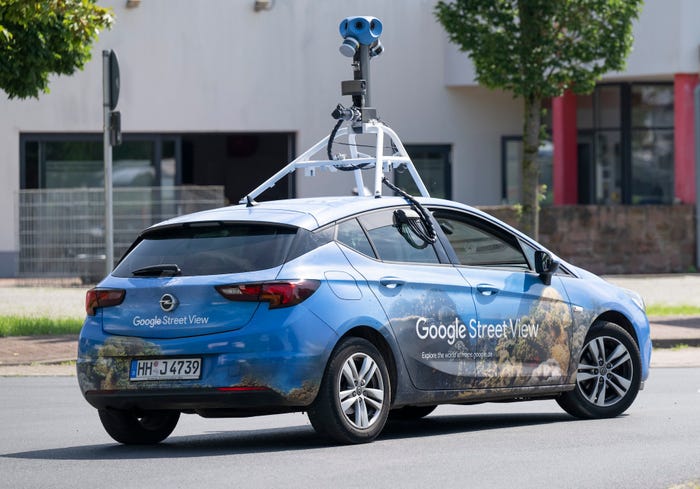 A blue Google Street View car, featuring a multi-lens camera affixed to the top, drives down a street in Germany. The car features an image of a coral reef, and the words "Explore the world at maps.google.de."