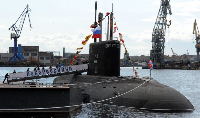 A black submarine sits in the water next to a dock. Sailors walk up a ramp to get into the submarine.