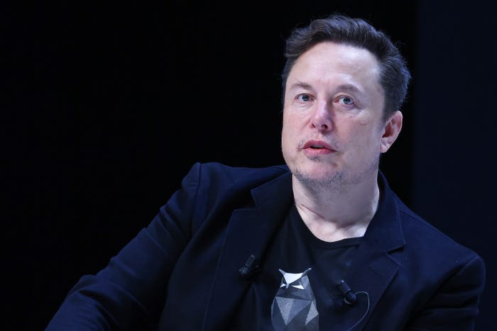 Elon Musk, wearing a small microphone on the lapel of his blazer, against a black background.