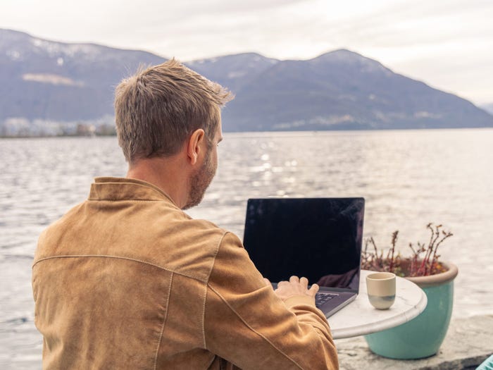 A man using a laptop at a table by a lake with mountains in the background.