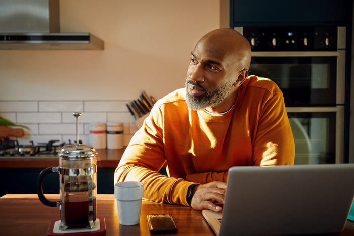 A bearded man sits in front of their computer in a kitchen and looks off to the side.